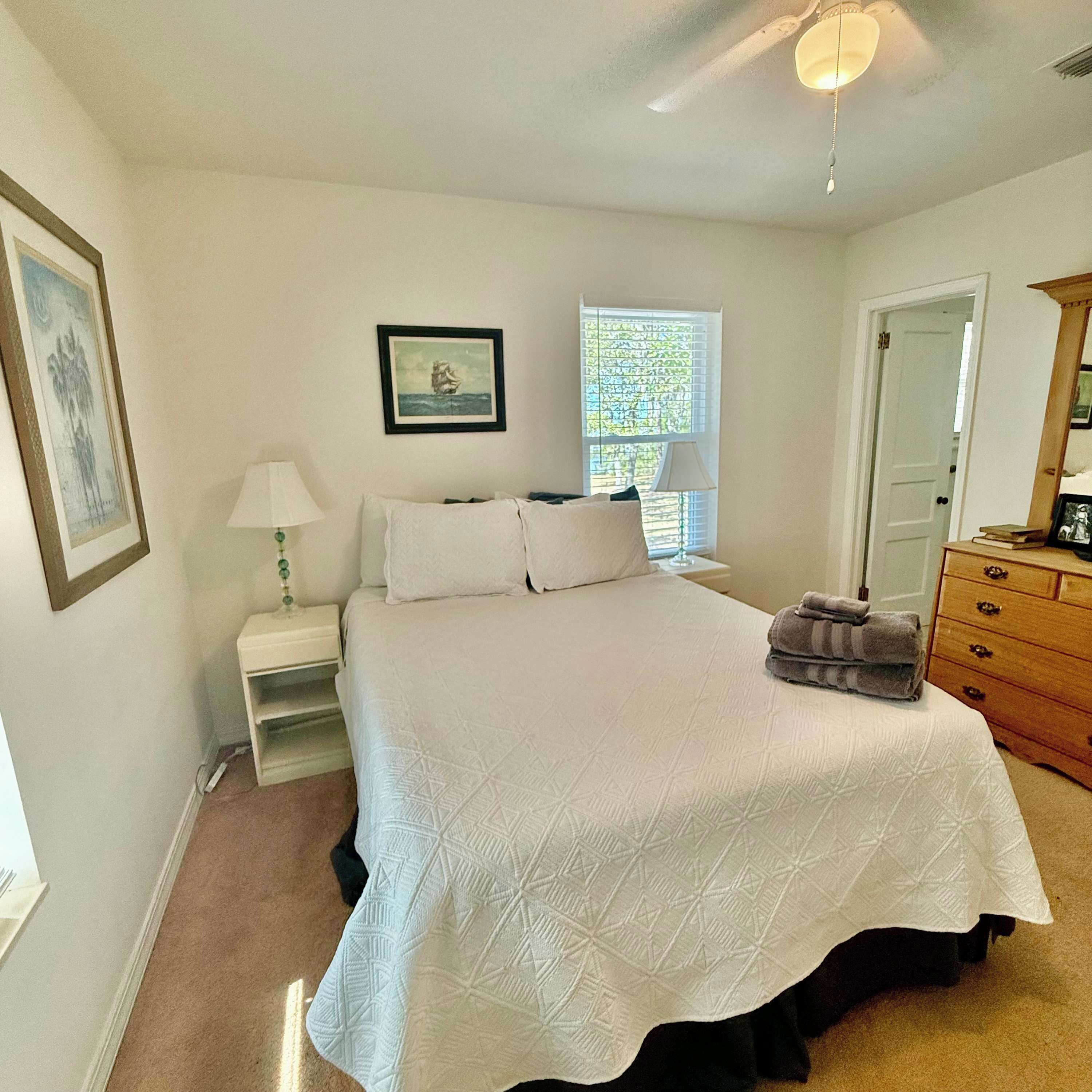 A neatly arranged bedroom with a large bed, a bedside table with a lamp, a window with blinds, and a door.
