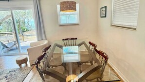 Een eetruimte met een tafel met glazen blad, omringd door houten stoelen, een geweven hanglamp en een uitzicht op het landschap door een groot raam.