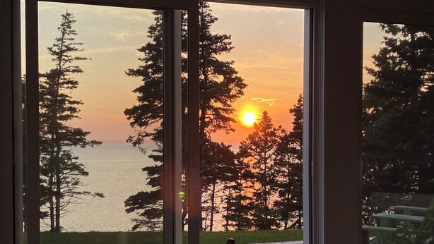 Ein Zimmer mit großem Fenster, das Blick auf einen Sonnenuntergang, eine Holzbank sowie das Meer und Bäume bietet.