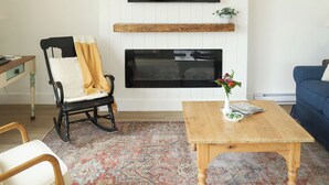 Ein Wohnzimmer mit Kamin, Wand-Fernseher, Schaukelstuhl, Couchtisch und einem gemusterten Teppich.