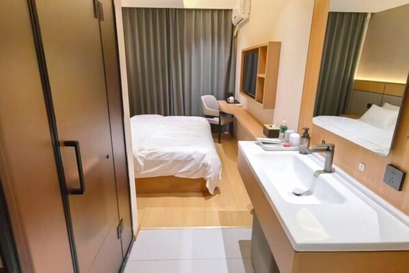 Habitación de hotel pequeña con una cama individual, un escritorio con silla, un espejo grande y un lavamanos blanco.