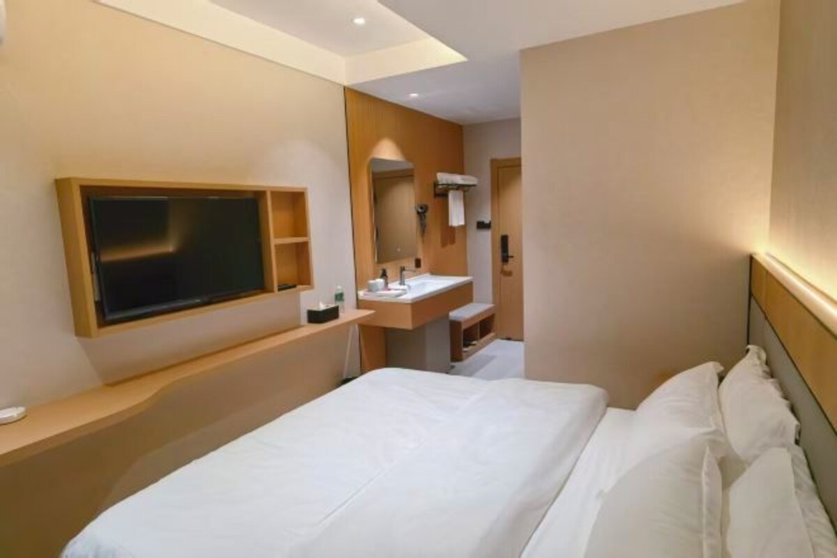 Una habitación de hotel moderna con una cama grande, un televisor montado en la pared, un baño con lavamanos y espejo, y un estante de madera.