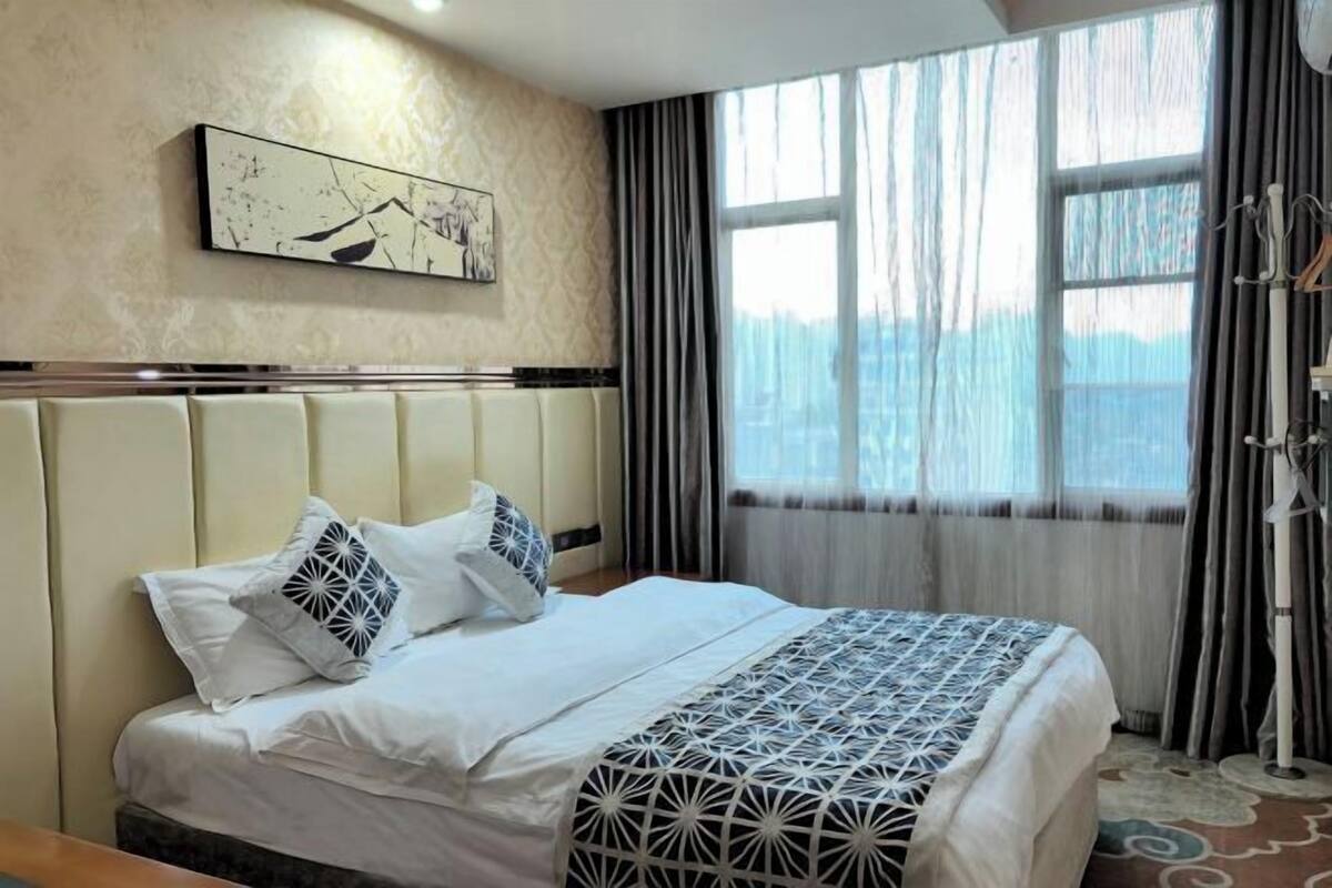 A hotel room with a bed, a headboard, a window with curtains, and a wall-mounted artwork.