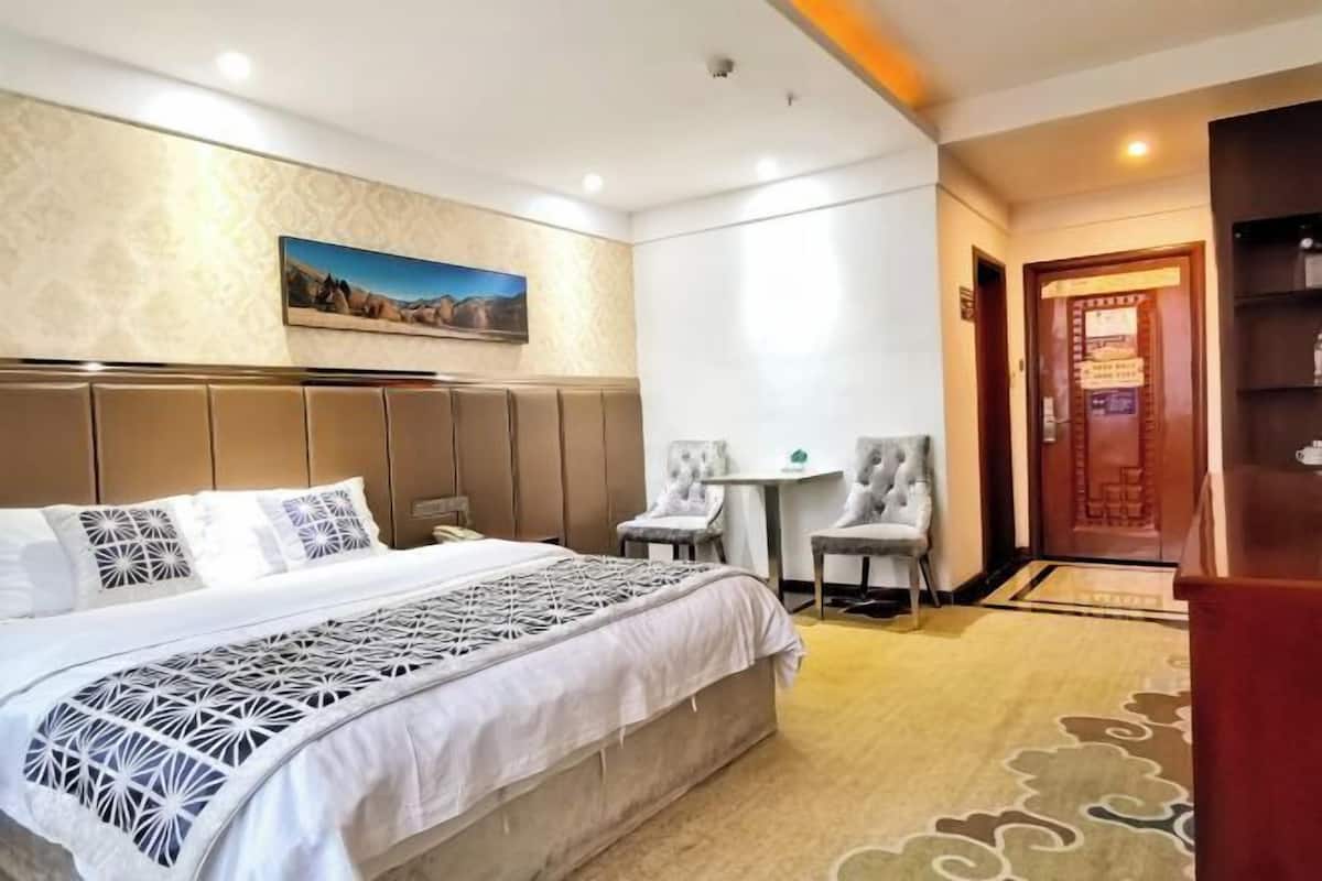 A hotel room with a large bed, a desk, two chairs, a painting on the wall, and a door with a sign.