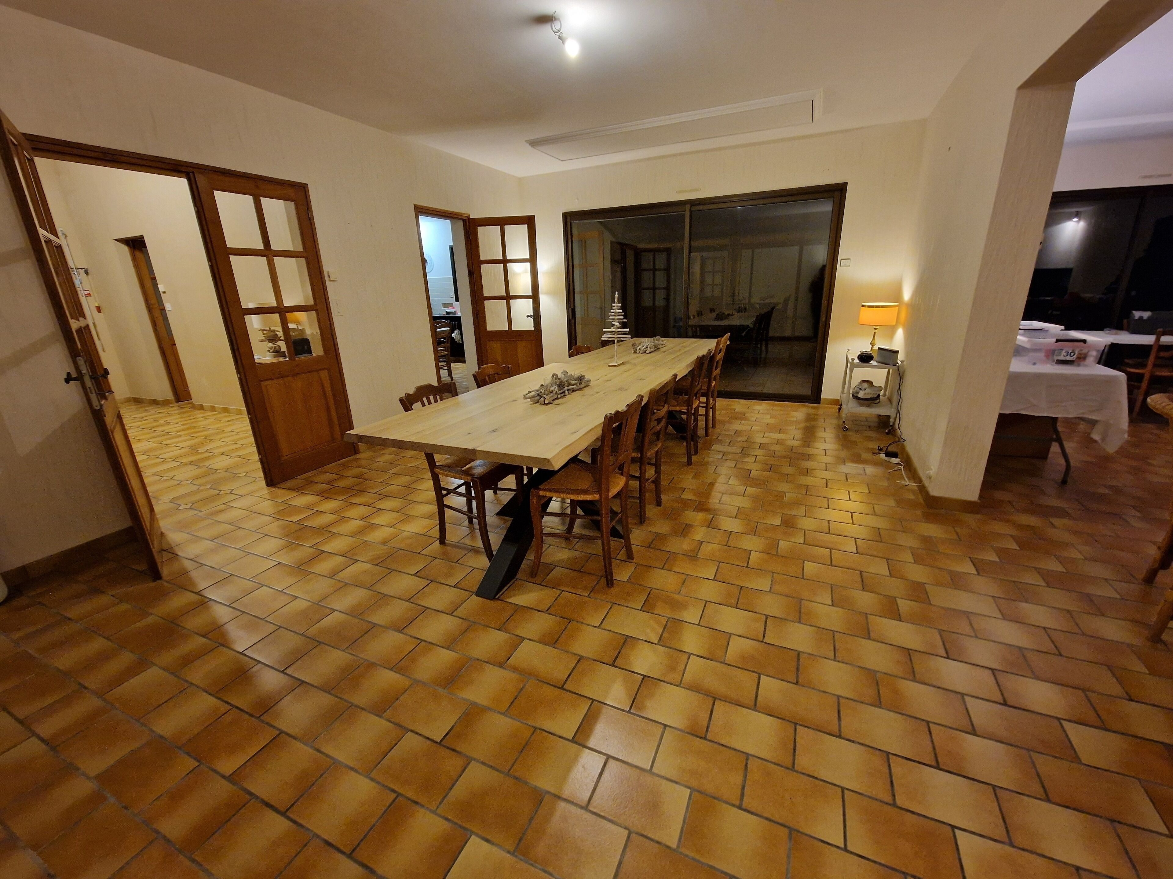 Un espace repas avec une longue table et des chaises en bois, un sol carrelé et une porte donnant sur une autre pièce.