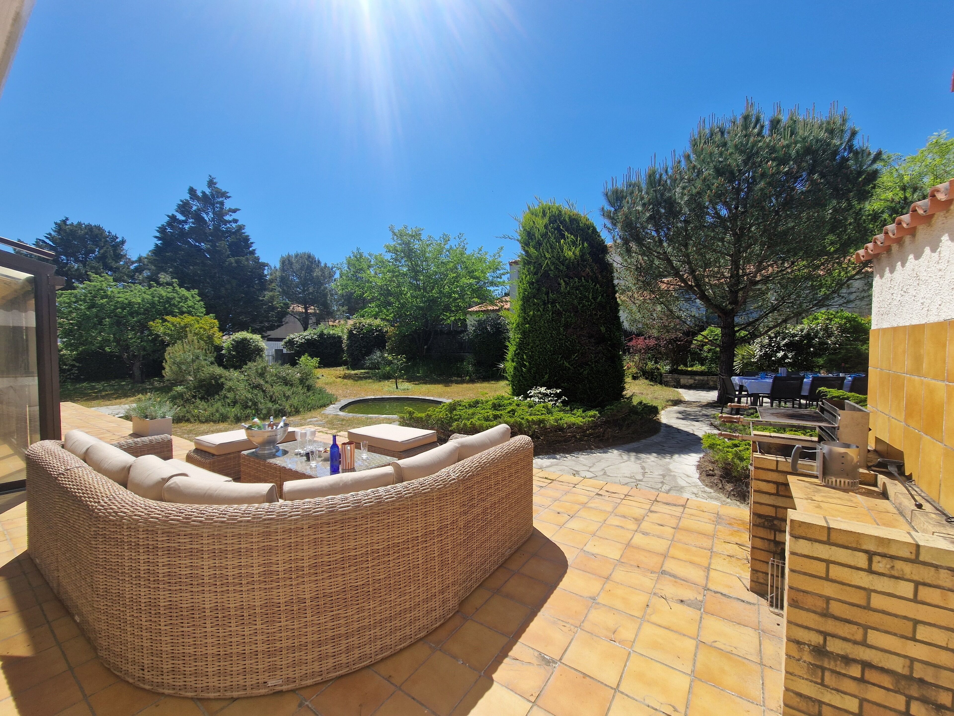 Une terrasse avec un canapé en osier, une petite table avec des verres et une vue sur un jardin agrémenté d’un bassin et d’arbres.