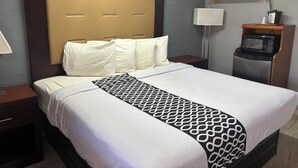 A hotel room with a large bed, a bedside table with a lamp, a microwave, and a small wooden side table with a tissue box. - Budget Inn (Nocona)