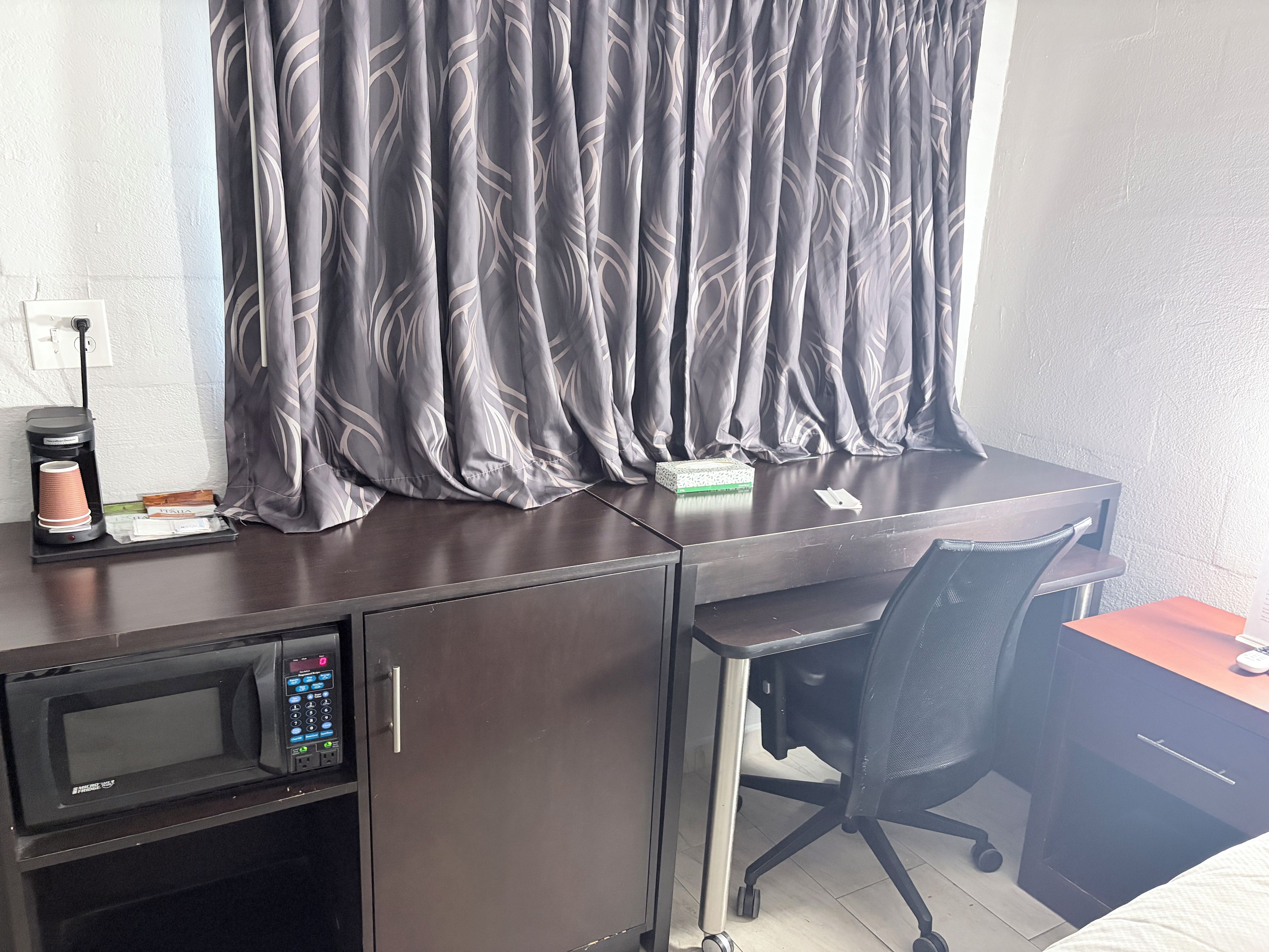 A hotel room with a desk, a chair, a microwave, a coffee maker, and a curtain.