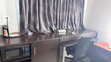 A hotel room with a desk, a chair, a microwave, a coffee maker, and a curtain.