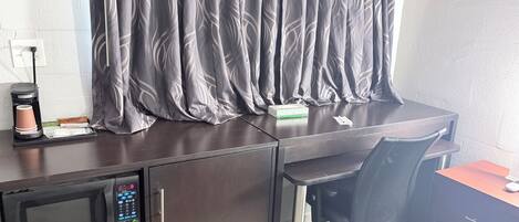 A hotel room with a desk, a chair, a microwave, a coffee maker, and a curtain.