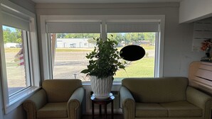 A living room with two beige armchairs, a large potted plant, and a view of a parking lot through large windows. - Budget Inn (Nocona)