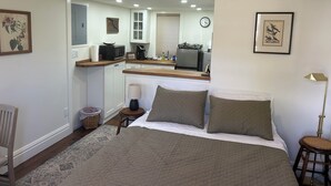 Een slaapkamer met een bed, nachtkastje, lamp en een stoel. Er is een kitchenette met een magnetron, koffiezetapparaat en een koelkast.