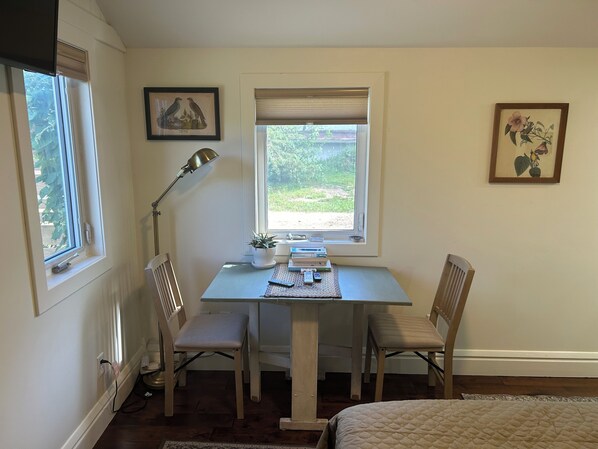 A small, neatly arranged room with a table, two chairs, a desk lamp, a potted plant, and framed pictures on the walls.
