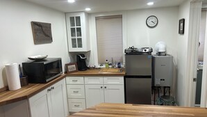 Een kleine keuken met witte kastjes, een houten aanrecht, een magnetron, een koffiezetapparaat, een koelkast en een klok aan de muur.