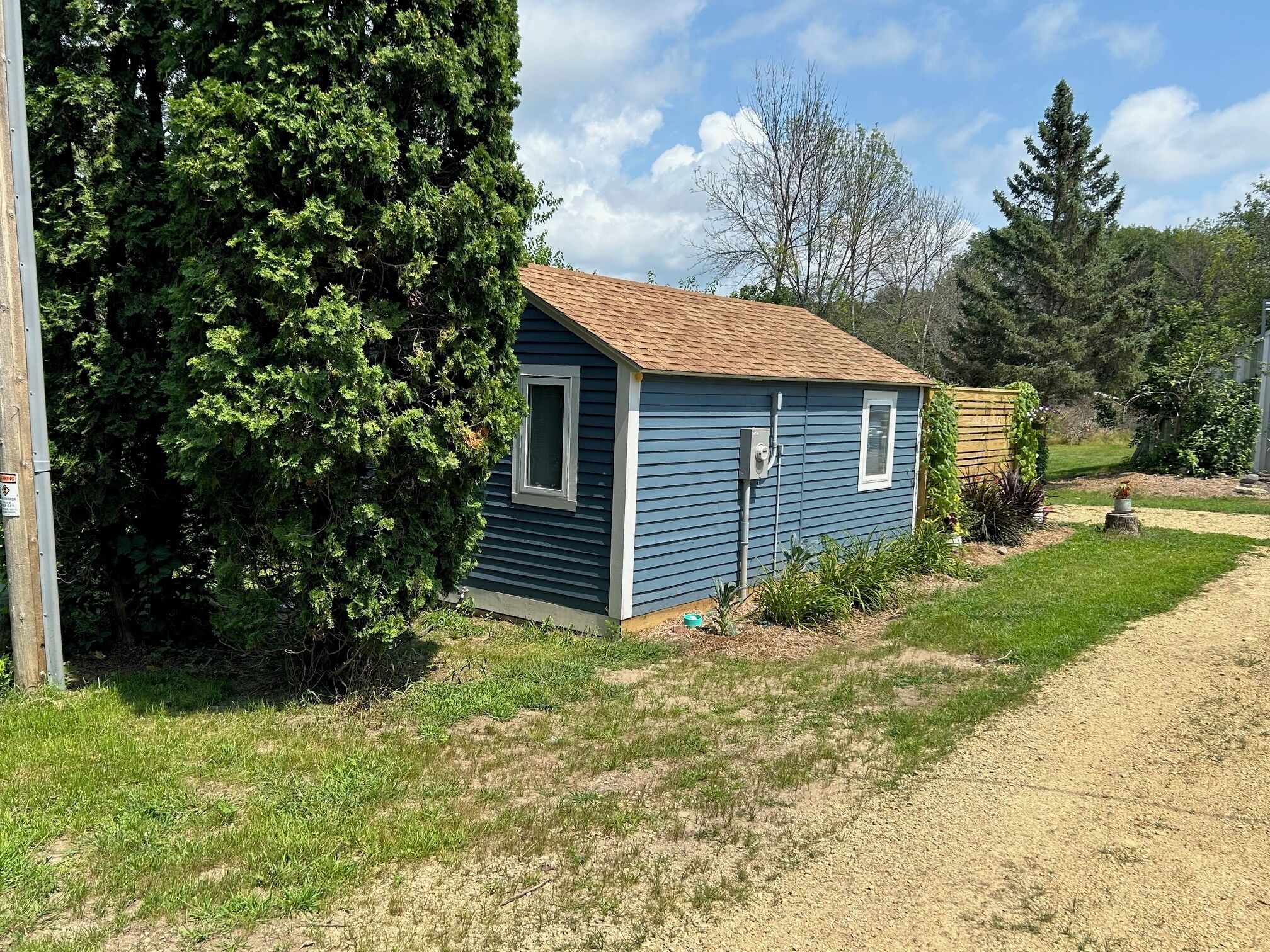 A small, single-story house with a brown roof and blue siding, surrounded by a grassy yard and trees.