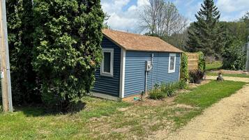 A small, single-story house with a brown roof and blue siding, surrounded by a grassy yard and trees.