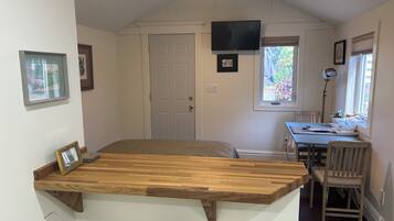Un dormitorio compacto con una cama de madera, un televisor montado en la pared, un escritorio pequeño con una silla y un ventanal con vistas a la vegetación.