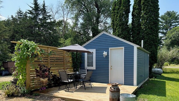 Een kleine blauwe schuur met een witte deur, een terras met een tafel en stoelen, en een houten omheining met planten.