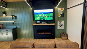 A living room with a TV on a black wall, a fireplace, a brown leather sofa, and a green wall with shelves. - Pepper Jack Shack: Your Cozy Mountain Retreat (Newport)