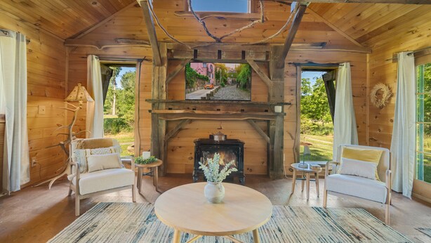 A cozy wooden cabin interior with a fireplace, two armchairs, a round table, and a view of a street through a window.