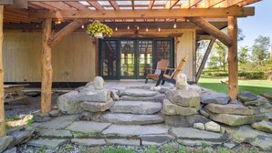 A wooden pergola with string lights and hanging flowers, a stone pathway leading to a wooden bench, and a wooden door with a view of greenery outside.