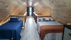 A narrow attic bedroom with two beds, wooden railings, and a window with curtains. - Bear Cabin: Charming 2-bedroom cabin with WiFi, AC in marvelous Sebec (Sebec)