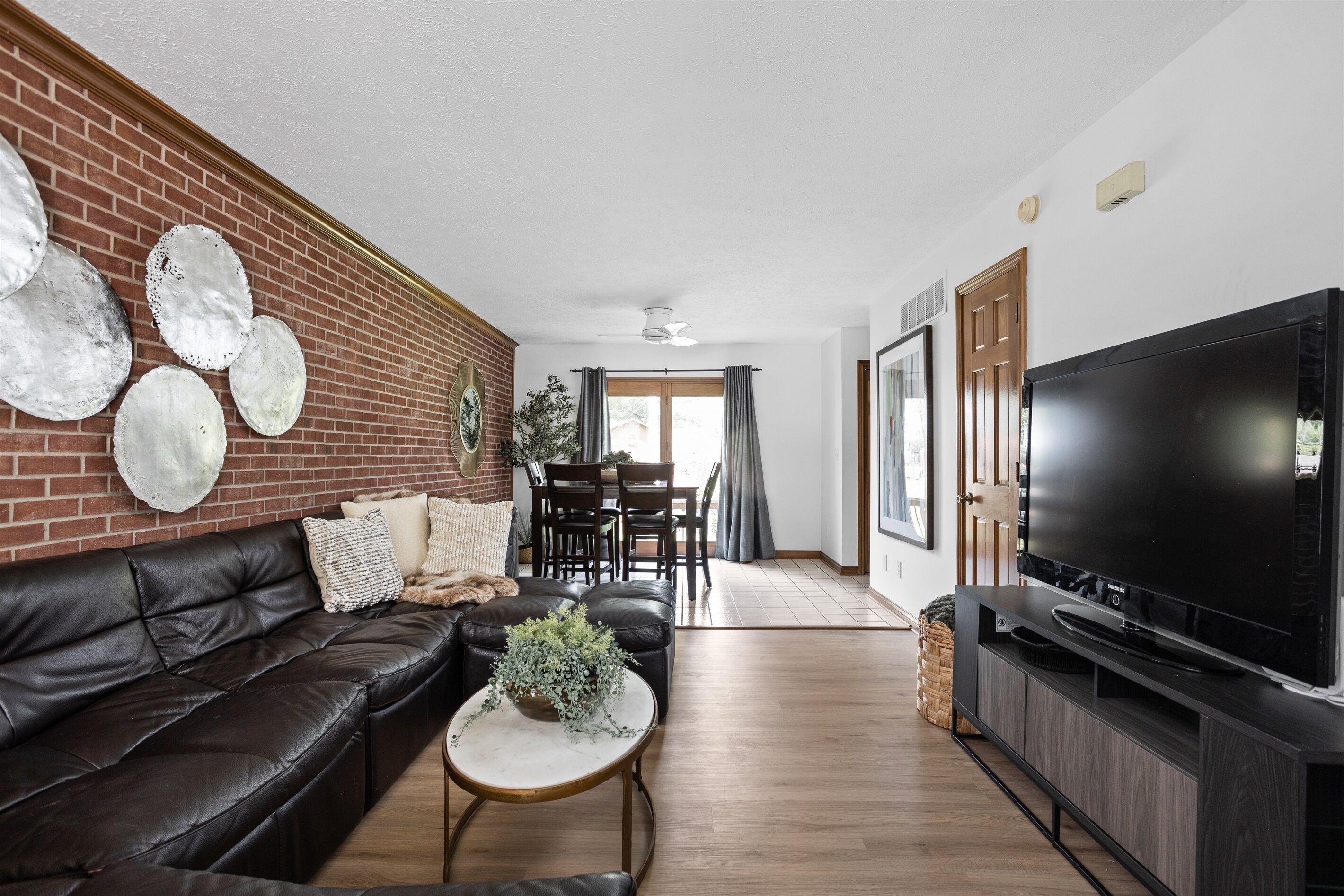 Un salon avec un canapé en cuir noir, une table basse agrémentée d’une plante, un téléviseur posé sur un meuble et un mur en briques orné d’assiettes décoratives.