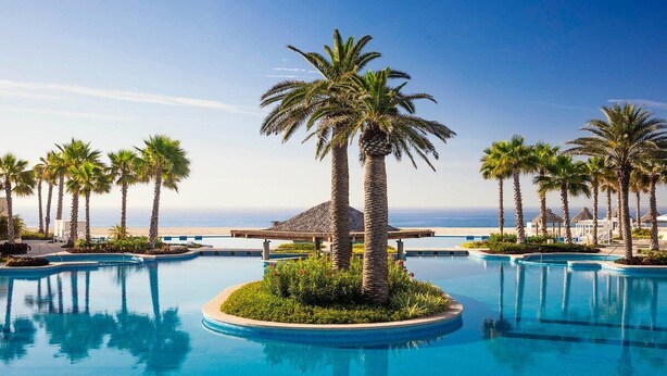 Área de piscina em resort com palmeiras, estrutura com telhado de palha e vista para o oceano.