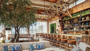 Um espaço de bar acolhedor, com mobiliário de madeira, uma árvore grande e estantes com várias garrafas e decorações.