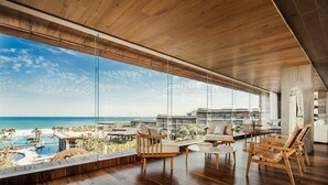 Interior moderno com uma grande janela de vidro com vista para uma piscina e praia, piso de madeira e móveis contemporâneos.