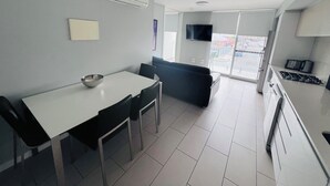 A modern kitchen with a dining area, white cabinetry, a flat-screen TV, and a large window offering a view of the street. - One Bedroom Apartment (Hamilton)