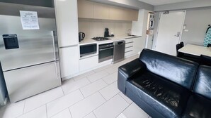 A modern kitchen with stainless steel appliances, a built-in oven, and a dishwasher. There is a black leather sofa in the living area. - One Bedroom Apartment (Hamilton)