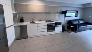 A modern kitchen with white cabinets, a built-in oven, and a microwave. There is a black sofa and a TV mounted on the wall. - One Bedroom Apartment (Hamilton)