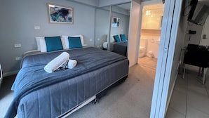A hotel room with a bed, a ceiling fan, a painting, a bathroom, and a TV mounted on the wall. - One Bedroom Apartment (Hamilton)