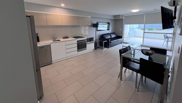 A modern kitchen with white cabinets, a dining area with a glass table, and a black sofa. - One Bedroom Apartment (Hamilton)
