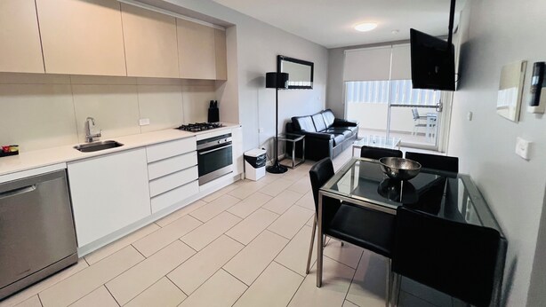 A modern kitchen with white cabinets, a gas stove, and a dishwasher. There is a dining area with a glass table and black chairs. - One Bedroom Apartment (Hamilton)