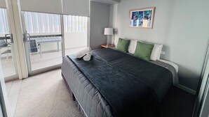 A hotel room with a bed, a nightstand, a lamp, a painting, and a sliding door to a balcony. - One Bedroom Apartment (Hamilton)