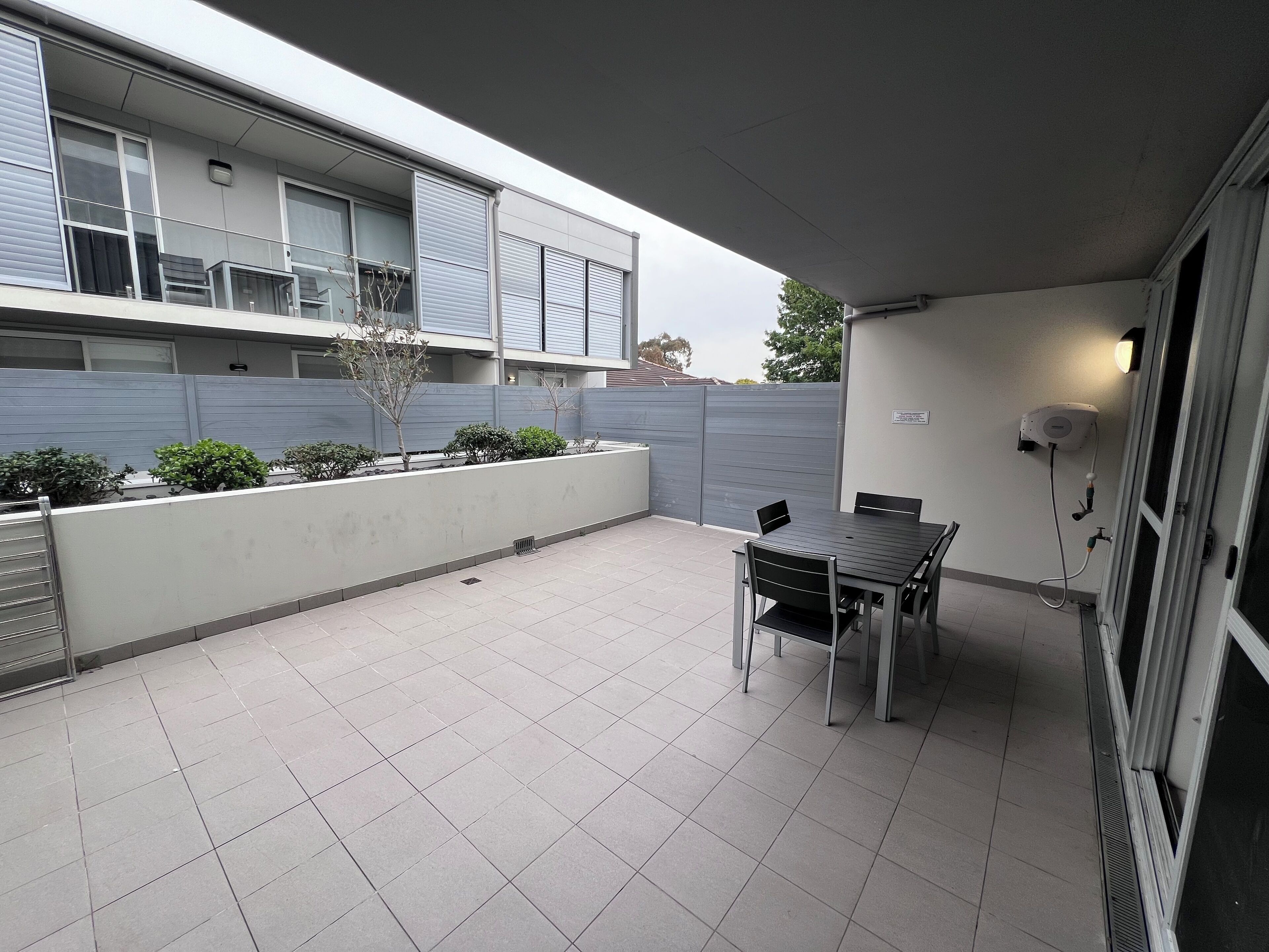 A covered patio with a table and chairs, a grey fence, and a building in the background.