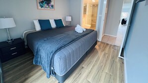 A modern bedroom with a bed, bedside tables, a ceiling fan, and a bathroom visible through an open door. - Two Bedroom Apartment (Hamilton)