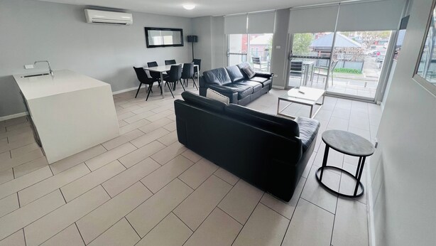 A modern living room with a black sofa, a white coffee table, and a dining area with chairs and a table. - Two Bedroom Apartment (Hamilton)