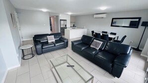 A modern living room with two black leather sofas, a glass coffee table, and a dining area with a white table and black chairs. - Two Bedroom Apartment (Hamilton)