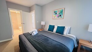 A hotel room with a large bed, a bedside table, a nightstand, a painting on the wall, and a door leading to a bathroom. - Two Bedroom Apartment (Hamilton)