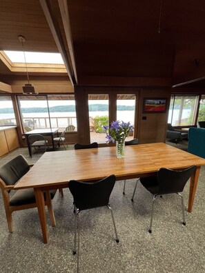 A conference room with a large wooden table, black chairs, and a vase with purple flowers. Large windows offer views of a lake and hills. - Spacious 2 bedroom home with 2 bedroom cabin on the water at Hood Canal. (Belfair)