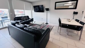 A modern living room with a black leather sofa, a glass coffee table, a dining table with chairs, a flat-screen TV mounted on the wall, and a large window with a view of buildings outside.
