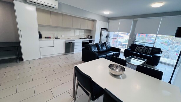 A modern kitchen with white cabinets, a dining area with a white table and black chairs, and a black leather sofa.
