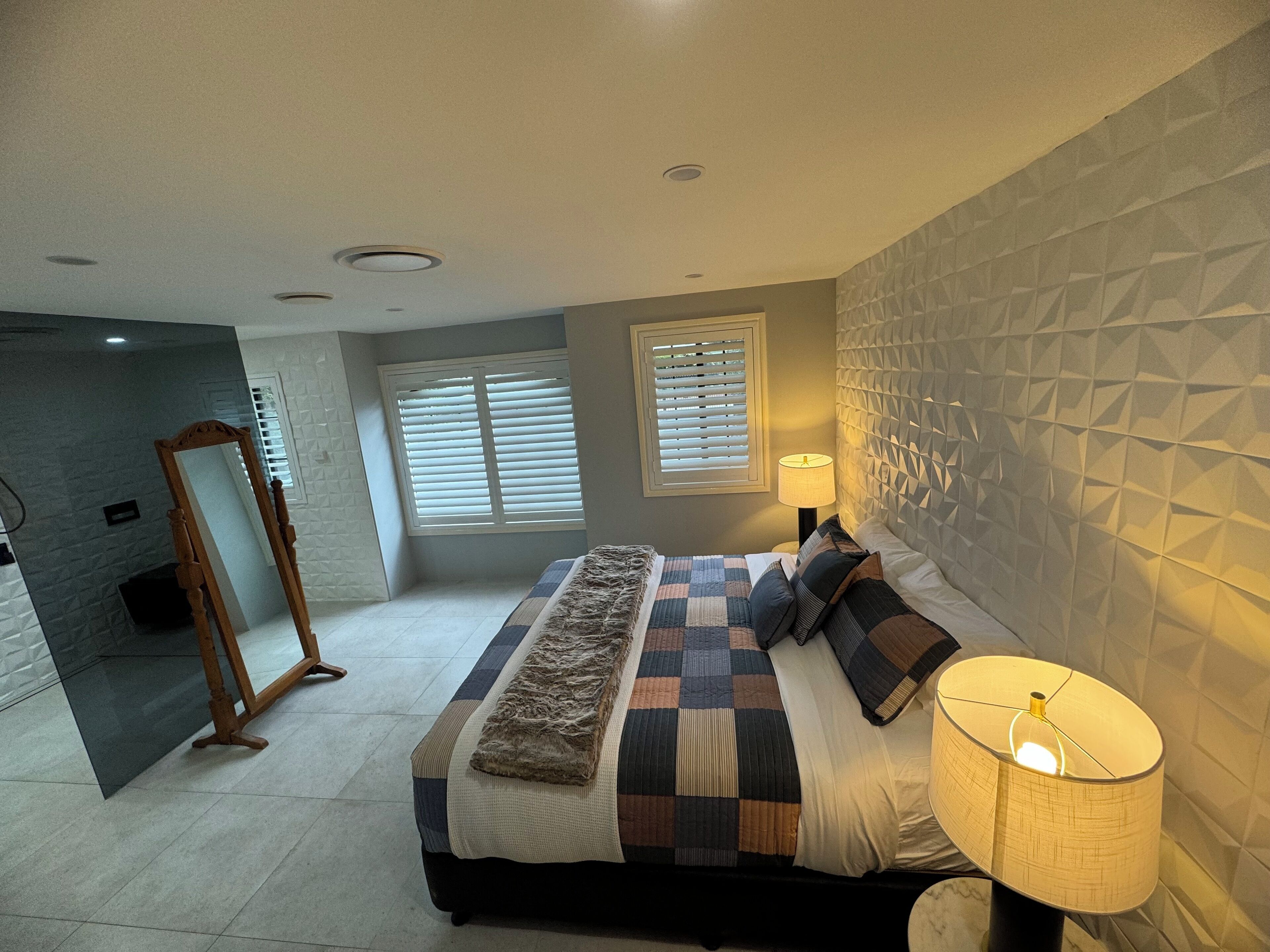 Une chambre à coucher avec un grand lit, une lampe de chevet, un miroir et une fenêtre avec des stores.