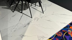 Zona pranzo con tavolo in legno e sedie nere, pavimento in marmo bianco e parete scura.