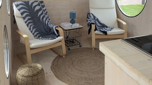 A compact, modern room with wooden walls and a large window. It includes two chairs, a small table with a lamp, and a kitchen area. - Hébergement Insolite Dans L'esprit Tinyhouse: la Tiny Bulle Pour Deux Personnes (Vignot)