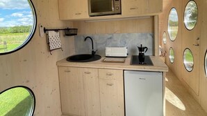 A compact kitchen with wooden cabinets, a microwave, a sink, a toaster, and a small refrigerator. There are windows on the walls, allowing natural light and views of the outdoors. - Hébergement Insolite Dans L'esprit Tinyhouse: la Tiny Bulle Pour Deux Personnes (Vignot)