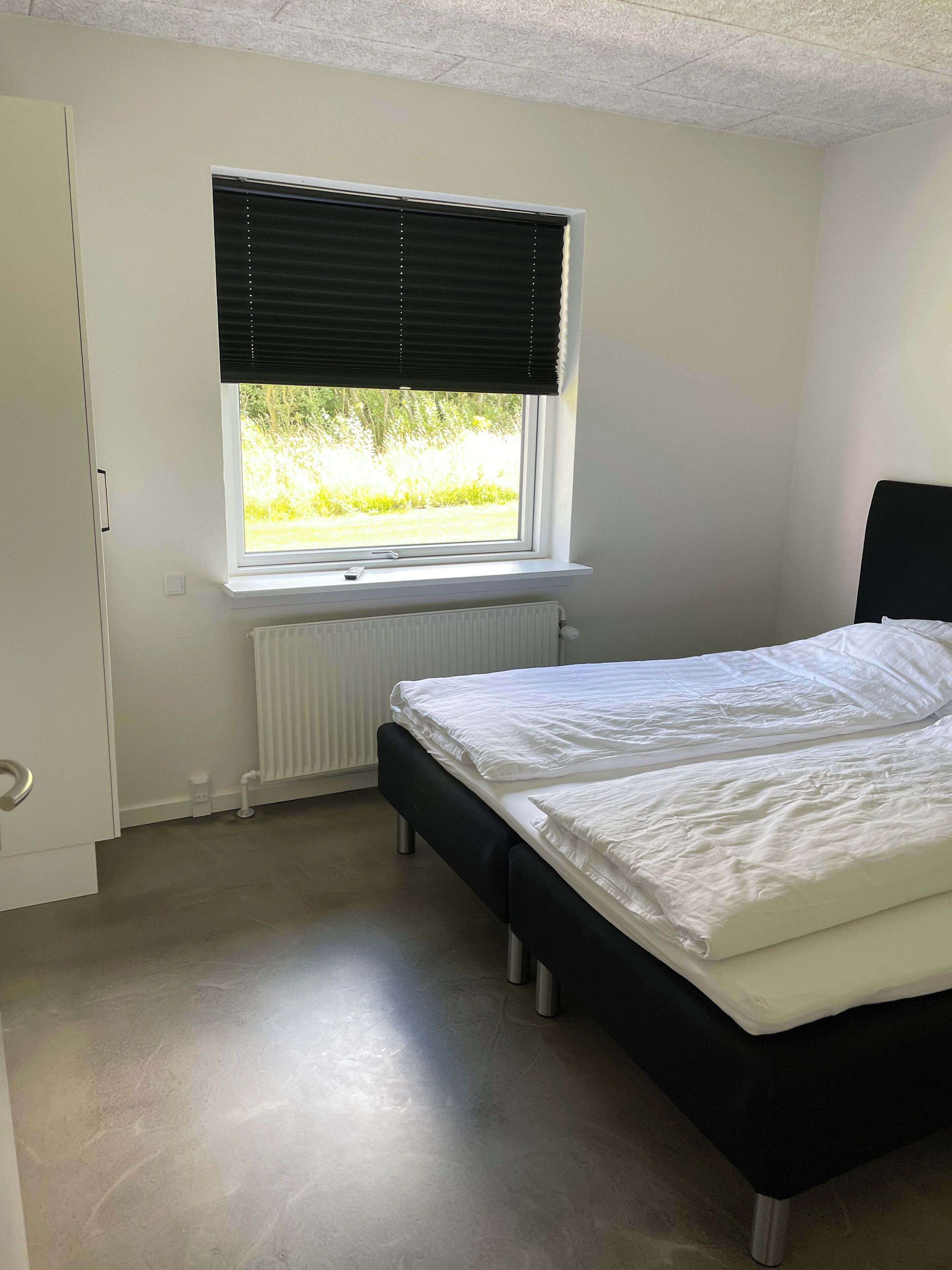 Ein Schlafzimmer mit Bett, Heizkörper, Fenster mit Jalousien und einer Tür.